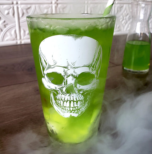 Egads Shop clear 16 ounce tumbler pint drinking glass with white screen print image of skull. Green, bubbling liquid fills the glass as white fog or smoke fills the foreground.